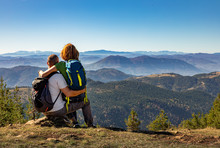 Rear View Of Hikers Couple With Backpack Sitting On Top Of The Mountain And Enjoying The View During The Day.