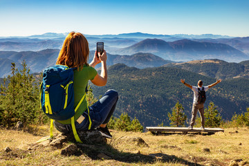 Wall Mural - hikers couple with backpack on top of the mountain, woman photographing her husband with phone.