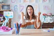 © Krakenimages.com - Young beautiful teacher woman wearing sweater and glasses sitting on desk at kindergarten very happy and excited doing winner gesture with arms raised, smiling and screaming for success.