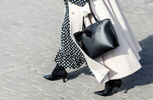 Woman In Autumn Or Spring In A Beige Coat And A Black Dress With White Polka Dots. Fashionable Bag Close-up In Female Hands.Girl Walks In The City Outdoors. Stylish Modern And Feminine Image, Style.