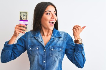 Wall Mural - Young beautiful woman holding dollars standing over isolated white background pointing and showing with thumb up to the side with happy face smiling