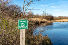 A Sign Marks The End Of The Trail, With A River, Grass And Trees In The Background. Concepts Of Outdoor Recreation, Hiking, And Parks