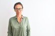 Middle age woman wearing green shirt and glasses standing over isolated white background with serious expression on face. Simple and natural looking at the camera.
