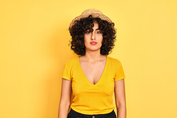 Sticker - Young arab woman with curly hair wearing t-shirt and hat over isolated yellow background with serious expression on face. Simple and natural looking at the camera.