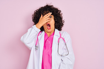 Sticker - Young arab doctor woman with curly hair wearing stethoscope over isolated pink background peeking in shock covering face and eyes with hand, looking through fingers with embarrassed expression.