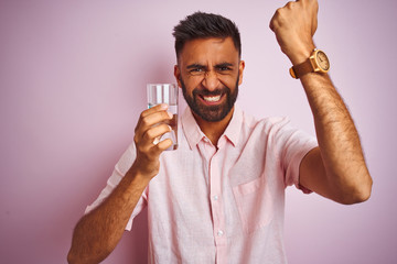 Wall Mural - Young handsome indian man dinking glass of water over isolated pink background annoyed and frustrated shouting with anger, crazy and yelling with raised hand, anger concept