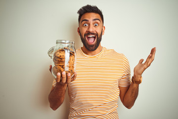 Poster - Young indian man holding jar of cookies standing over isolated white background very happy and excited, winner expression celebrating victory screaming with big smile and raised hands