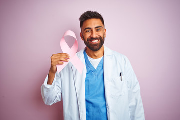 Wall Mural - Young indian doctor man holding cancer ribbon standing over isolated pink background with a happy face standing and smiling with a confident smile showing teeth