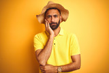 Fototapeta  - Young indian man on vacation wearing summer hat standing over isolated yellow background thinking looking tired and bored with depression problems with crossed arms.