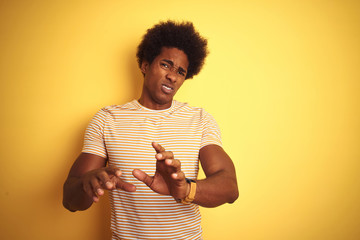 Sticker - American man with afro hair wearing striped t-shirt standing over isolated yellow background disgusted expression, displeased and fearful doing disgust face because aversion reaction. 