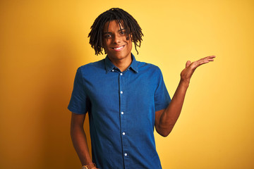 Afro man with dreadlocks wearing casual denim shirt standing over isolated yellow background smiling cheerful presenting and pointing with palm of hand looking at the camera.