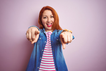 Sticker - Beautiful redhead woman wearing denim shirt and striped t-shirt over isolated pink background pointing to you and the camera with fingers, smiling positive and cheerful