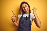 Young beautiful hairdresser woman holding scissors over yellow isolated background screaming proud and celebrating victory and success very excited, cheering emotion