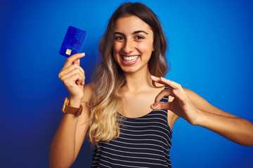 Poster - Young beautiful woman holding credit card over blue isolated background doing ok sign with fingers, excellent symbol