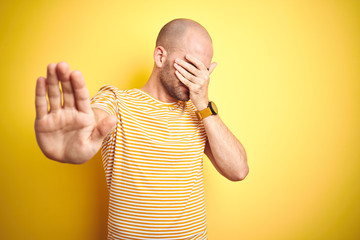 Wall Mural - Young bald man with beard wearing casual striped t-shirt over yellow isolated background covering eyes with hands and doing stop gesture with sad and fear expression. Embarrassed and negative concept.