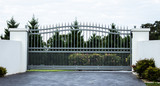 Fototapeta  - Grey metal wrought iron driveway property entrance gates set in white concrete brick fence, garden trees in background