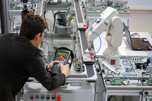 Man Is Holding Teach Panel To Control A Robotic Arm Which Is Integrated On Smart Factory Production Line. Industry 4.0 Automation Line Which Is Equipped With Sensors And Robotic Arm. Selective Focus.