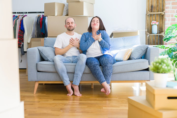 Canvas Print - Young couple sitting on the sofa arround cardboard boxes moving to a new house smiling with hands on chest with closed eyes and grateful gesture on face. Health concept.