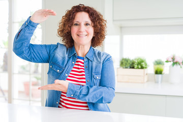 Poster - Middle age senior woman with curly hair wearing denim jacket at home gesturing with hands showing big and large size sign, measure symbol. Smiling looking at the camera. Measuring concept.