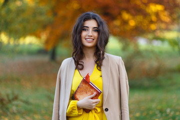 A beautiful smiling brunette woman with a curly hairstyle in the park is reading a book. Fashionable autumn clothes in orange mustard tones, autumn blur colors backgrounds.