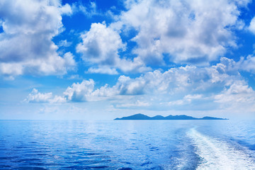 Wall Mural - White cumulus clouds in blue sky over sea landscape, many clouds above ocean water panorama, ship trail, island on horizon line, beautiful tropical sunny summer seascape panoramic view, cloudy weather