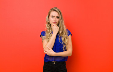 young blonde woman looking serious, confused, uncertain and thoughtful, doubting among options or choices against red wall