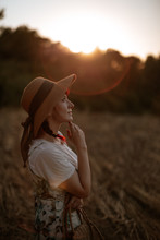 Side View Of Female In Retro Dress And Hat Walking In Field Towards Sunset Sky While Looking Away