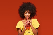 Dreamy Young African American Lady With Curly Hair Holding Red Jar With Straw And Enjoying Beverage On Red Background