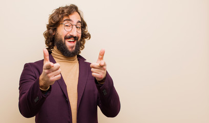 young bearded crazy man smiling with a positive, successful, happy attitude pointing to the camera, making gun sign with hands against flat color wall