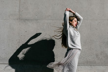 Side View Of Young Female Model In Stylish Warm Sweater And Skirt Closing Eyes Raising Elbow While Standing Against Gray Wall On City Street