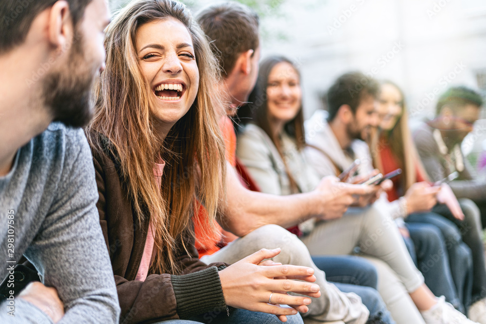 Group of trendy young people chatting together sitting on a bench outdoors. Students having fun together. Focus on a blonde girl smiling with open mouth - obrazy, fototapety, plakaty 