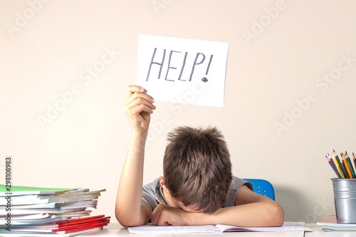 Fotomural Sad tired frustrated boy sitting at the table with many books and holding paper with word Help