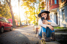 Beautiful Stylish Woman Sitting On Street At Sunny Day And Using Mobile Phone With Headphones