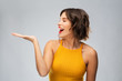 emotions and people concept - happy smiling young woman in mustard yellow top holding something on empty hand over grey background