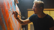 © Gorodenkoff - Talented Artist Working on Abstract Painting, Dips Brush in Paint and Starts Drawing Daringly Emotional Modern Art. Dark Creative Studio Large Canvas Stands on Easel Illuminated