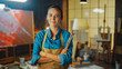 © Gorodenkoff - Professional Young Female Artist Dirty with Paint, Wearing Apron, Arms Crossed while Holding Brushes, Looks at the Camera with a Smile. Authentic Creative Studio with Large Canvas. Face Portrait