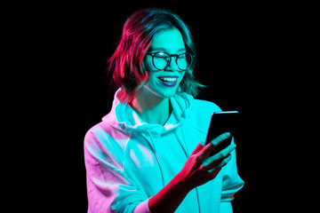 technology and people concept - happy young woman wearing hoodie and using smartphone in neon lights over black background