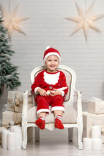 Happy Little Smiling Boy In Santa Claus Costume Sits On Armchair Near Christmas Tree And Holds White Candle In Hands