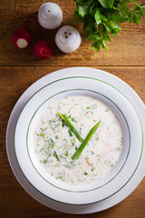 Wall Mural - Classic cold soup Okroshka made of vegetables, boiled potatoes, eggs, and meat (beef, sausages or ham) in white bowl, wooden table. Overhead, vertical image