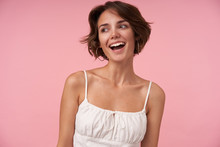 Studio Shot Of Charming Young Female With Casual Hairstyle Wearing White Top While Standing Over Pink Background, Looking Aside Cheerfully And Smiling Widely, Being In Nice Mood