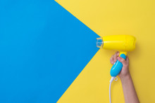 The Right Hand Holds A Hair Dryer Simulating The Flow Of Air On A Yellow And Blue Background.