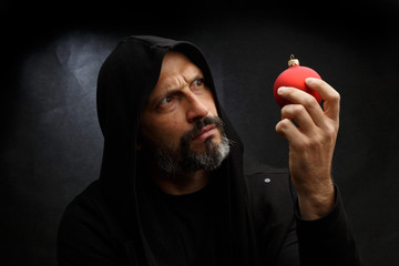 Wall Mural - Portrait of a bald man with a beard in a black hood witha red christmas ball on a dirty gray background. Young Santa concept.