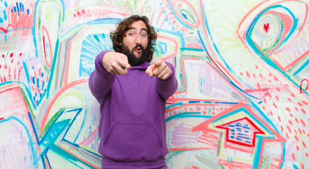 young bearded crazy man feeling happy and confident, pointing to camera with both hands and laughing, choosing you against graffiti wall