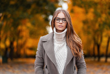 Autumn Portrait Of An Attractive Young Woman In Stylish Glasses In A Knitted Fashionable White Sweater In An Elegant Coat In A Park On A Background Of Trees With Orange Leaves.Girl Walks In The Forest