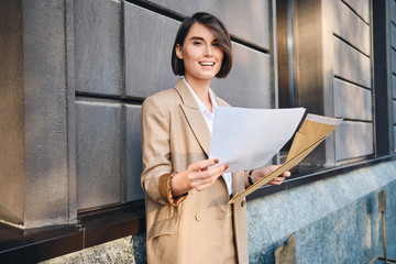 Young joyful businesswoman happily looking in camera working with papers outdoor