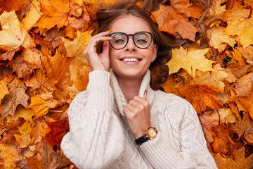 Young model of a joyful woman lies in orange leaves and straightens stylish glasses. Pretty positive girl hipster in a beautiful smile in a knitted sweater is resting on an outdoor. View from above.