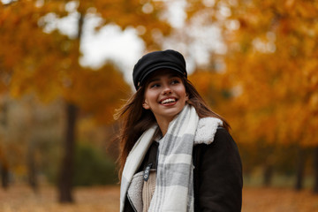 Model of a positive young woman in a stylish hat in a brown jacket in a scarf for a walk in the autumn forest. Joyful girl with a cute smile is resting in the park on a background of golden foliage.