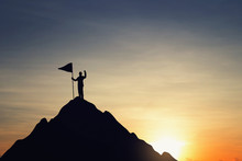 Silhouette Of Businessman Holding A Flag On Top Mountain, Sky And Sun Light Background. Business Success And Goal Concept.