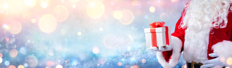 Wall Mural - santa claus holding a christmas present with snowy background and golden lights