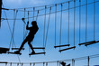 © Irin Fierce - Silhouette of young girl in helmet climbing on high rope course against blue sky. Wooden bars tied to ropes, part of obstacle course in adventure park